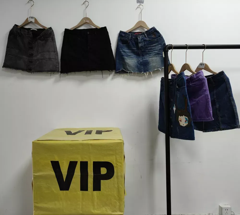 Denim skirts in various styles and colors, including black, blue, purple, and distressed designs, displayed on hangers with a VIP box in front, representing wholesale used clothing products from RossBoss.